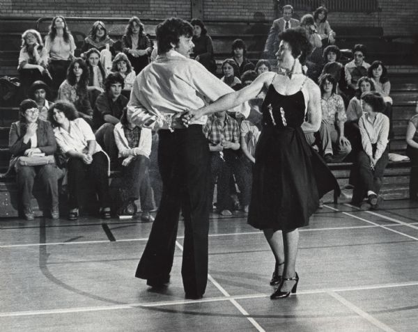 A man and a woman are dancing in a school gymnasium, while teenagers are watching from the bleachers. Caption reads: "The Mary D. Bradford Senior High School of Kenosha sponsored a 'Mini-Course' day Wednesday. Students participated in a number of courses offered by about 150 guest presenters. Disco dancing was one of the courses offered. Bella Bellegante, owner of Bellegante Dance Studuio, and dance instructor Wayne Beecher, both of Kenosha, demonstrated a disco step."