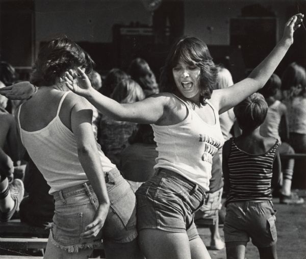 Two women dancing "the bump," wearing tank tops and jean shorts. One of the women's shirt reads "Handle With Care." Other people are in the background.