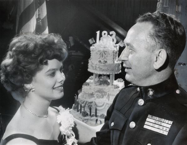 A man in uniform and a woman are standing together looking at each other; behind them is a cake on a table with frosting that reads: "U.S. Marines" and has the number "183" and American flags topping it. Caption reads: "The pleasure was all his at the annual birthday ball of the United States marine corps Monday night at the Memorial Center, Lt. Col. Anthony R. Frankiewicz, commanding officer of the 16th infantry reserve battalion, danced with Miss Sharon Shaw, 4035 N. 46th St. The reserve unit sponsored the ball here."