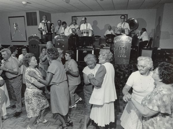 Several people, most of them women, dancing on a dance floor in front of a band on a stage. Caption reads: "CONCERTINA DANCING MUSIC — The woman outnumbered the men as dancers gathered for some old time music by the Milwaukee Cream City Concertina Club. The event was held at Chalet Danceland in Richfield."