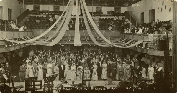 Elevated view from stage towards a large group of young people in formal attire standing in a decorated gymnasium. People are also up in the balcony. Caption reads: "Junior Prom, April 15, 1910, Antigo, Wis."