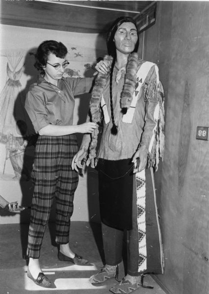 Dr. Joan Freeman adjusting the clothing on a mannequin of a Native American. Dr. Freeman was the first woman to receive a PhD. in Anthropology from UW-Madison, with a specialization in archaeology. She was also the first State Archaeologist. In 1960 she joined the Wisconsin Historical Society as Curator of Anthropology. 