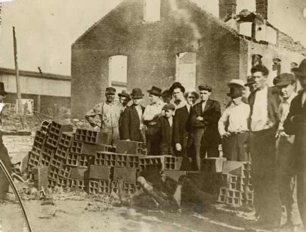 A group of white men are posing along a pile of cinder blocks. In front of the blocks, and partially obscured by shadows, is the body of a black man. Caption reads: "One of the victims of the East St. Louis, Ill., race riots.
The body can be seen lying on the ground in front of the pile of brick[sic]. This unfortunate man was dragged from the Cahokie Creek, pulled through the main streets, and finally killed by a shower of bullets rained upon him by the mob.
Exact number of dead in the East St. Louis race riots has not been ascertained but it is known that at least thirty have perished and the figures may mount as the sections burned by the mob are searched. Three hundred are known to have been injured. Governor Lowden of Illinois has arrived. 1200 Illinois Guardsmen are today patrolling the disturbed sections. Comparative quiet has been restored."