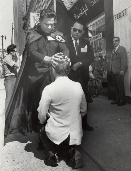 A caped man standing on a sidewalk is placing his hands on the head of a kneeling man, while another man in a suit and sunglasses is looking on. Caption reads: "A self-described warlock (male witch) in black flowing cape bestowed a blessing right here in Old Milwaukee Tuesday. Gene De Jean blessed the city and a number of passersby at N. 3rd St. and W. Wisconsin Ave. He was in town for a magician's convention."