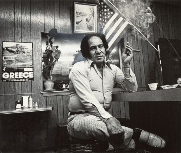 A man is sitting on a bar stool with his legs crossed, smoking a cigar. Behind him is an American flag, along with a poster or calendar for Greece. Caption reads: "Abu, the owner and operator of the Jerusalem of the Star restaurant."