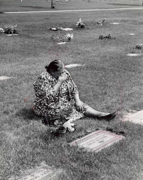 A woman is sitting and blowing her nose in front of a gravestone. A potted plant and a handbag are next to her. Caption reads: "Nine years after his death, Mrs. Rifa Zumot of Amman, Jordan, paid homage to her son at his grave at Graceland cemetery. The son, Abdullah, 28, died in May, 1954, of a ruptured appendix. He had been in Milwaukee for 15 months. Mrs. Zumot is staying with the John Krumbeck family, 5914 N. 70th St."