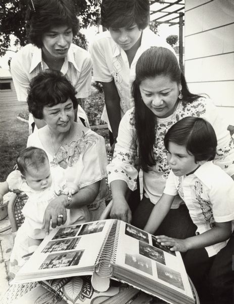 Six family members looking at a photograph album outdoors. Caption reads: "REMEMBERING. Norma Clemente and her family looked through the family album at her home in Oak Creek. Clockwise from left foreground were granddaughter Lisa, 5 months; sons Gene and Mark, daughter Dorothy and grandson Joey, 5."

An additional clipping from the newspaper reads: "When we were planning to come to America, lots of our friends in the Philippines tried to discourage us," Norma Clemente said. "They told us, 'Oh, life there is not so great, the people aren't friendly, your neighbors won't help you.'"
