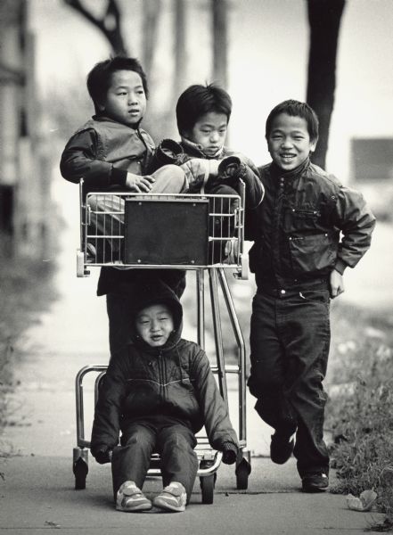 Four brothers posing in and next to a shopping cart. Caption reads: "Bee Vang ended up in the lower berth, and his brothers, Sam and John, rode on top as another brother, Meng, pushed them in a shopping cart they found near S. 10th and W. Bruce Sts."