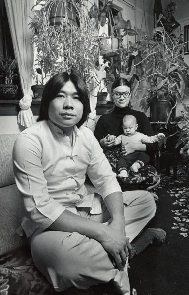 A man, woman, and infant posing in a room with several hanging plants. Caption reads: "Pamela Khavitone with Devin and her Laotian husband Mahn."