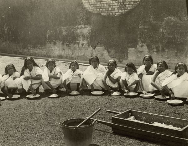 Ten girls are posing in a walled courtyard sitting on the ground, with plates of rice in front of them. In the foreground is a wooden box and a pail. Caption reads: "INDIA. Burma. Rangoon. Hindu girls of a mission school cleaning rice."