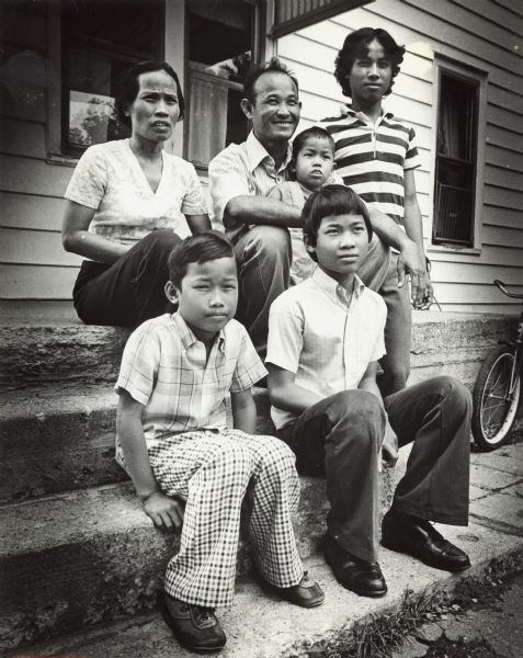 A Cambodian family of six posing on the front steps of their home. Caption reads: "In front of their home in Stoughton, the Phak family poses for a portrait. The family includes, back row from the left, Touch Prech, her husband Phon Phak, who is holding Sameth, and Roeun; front row from the left, Mon and Proeung."
