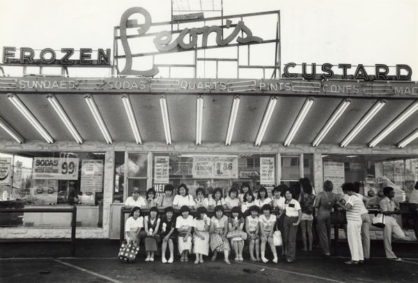 View across parking lot towards a group of Japanese woman posing in front of Leon's Frozen Custard. Caption reads: "Visiting Japanese students posed in front of Leon's frozen custard stand, one of the many stops the group made while touring Milwaukee."