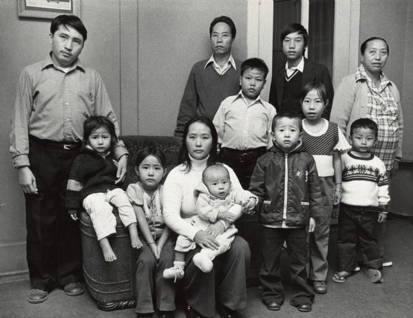 A family of twelve posing in a room; some are sitting and some are standing. Caption reads: "The Laotian family in Milwaukee posed for a portrait Monday. They are expecting word Tuesday on the treatment their relative, Mrs. Yeu Ly, who is suffering from bone marrow disease, should have. But since arriving here, the Laotians have found the stay in this country cold and confusing."