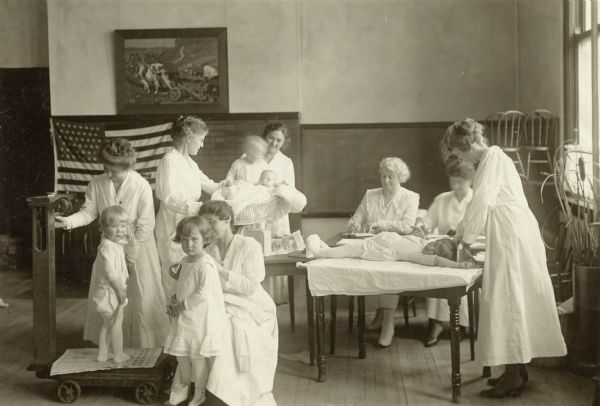 Seven women in white uniforms measuring five children, including one infant. Caption reads: "Lincoln School, Madison, Wis. Weighing and Measuring Campaign Summer 1918. Child Welfare Com. Women's Com. Dane Co. Council of Defense."