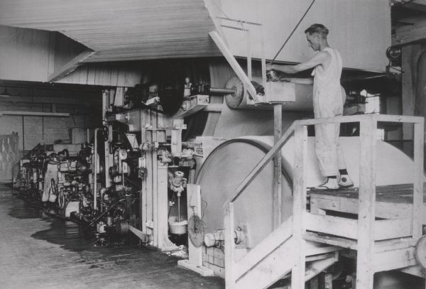 View towards a man in coveralls standing on a wooden platform operating a cellucotton processing machine.