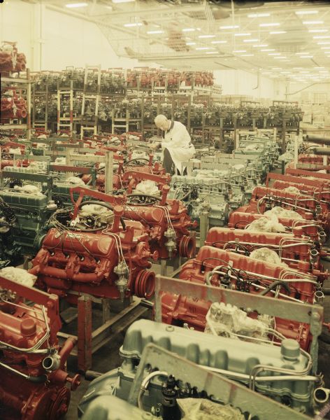 Interior view of the American Motors plant. A man in a lab coat is holding a clip board and examining rows of engines.