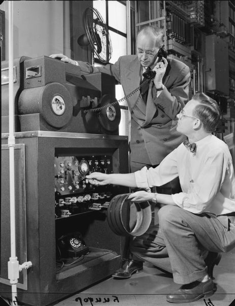 Stanley E. Bennett, a First Wisconsin National Bank cashier, listens to the handset while John T. Frymark, a Wisconsin Telephone Company central office foreman operates a monitor switch to test the Audichron. The Audichron was a machine located in the Wisconsin Telephone building at 722 N Broadway that reported the time via tape recordings when people called Broadway 1-4911 in Milwaukee. The service was sponsored by First Wisconsin National bank. In 1953, the machine fielded 3,166,305 calls.