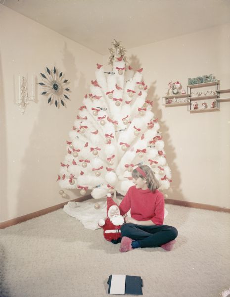 Portrait of a girl wearing a red top, black pants and pink slippers holding a Santa doll while sitting on the carpet in front of a white nylon Christmas tree at the William Driscoll home, 3765 S. 19th. The tree is decorated with red birds, blue birds, gold balls and red bows. A clock and a small shelf with holiday knickknacks decorate the walls.
