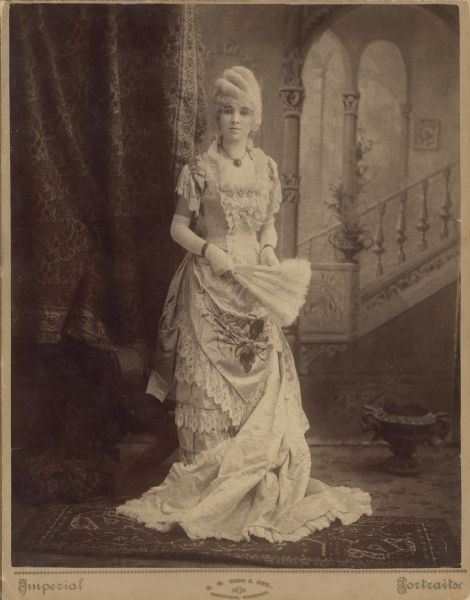 Full-length studio portrait in front of a painted backdrop of a young Adda Howie wearing a formal gown, gloves, and a wig. She is holding a feathered hand fan.