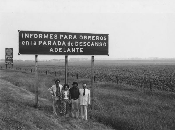 Four people are posing beneath a road sign that reads: "Informes para obreros en la parada descanco adelante" (Information for workers at the stop).