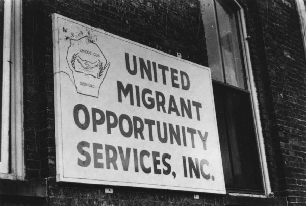 View of a United Migrant Opportunity Services sign. A logo consisting of the outline of the State of Wisconsin with two hands shaking are between laurels, and the phrase "Unidos con Dignidad" is at the top left.