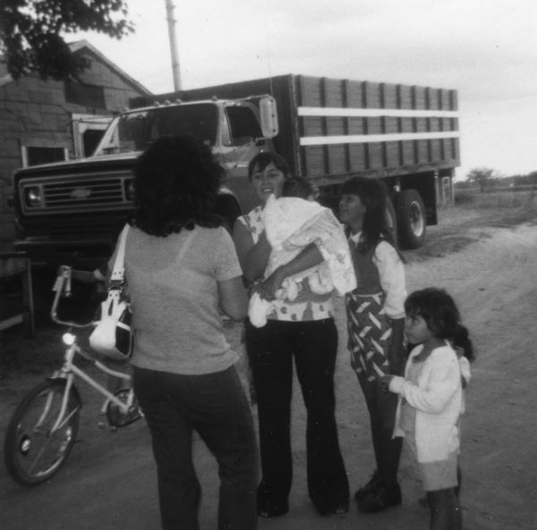 Two women and five children are standing and talking in a dirt road near a parked farm truck. One of the children, mostly obscured by a woman, is on a bicycle.