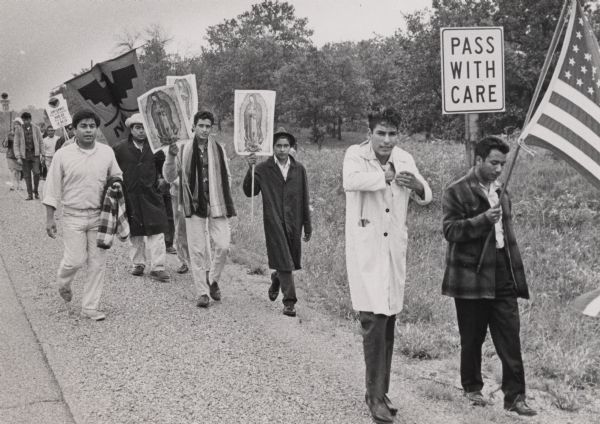 View of a group of migrant workers carrying signs during a march from Wautoma to Madison to promote workers' rights. Some of the people are carrying the image of Our Lady of Guadalupe, and one man is carrying a National Farm Workers Association banner.