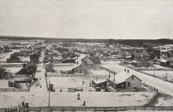 View overlooking Iron River in winter. Several residences, and yards with fences are in the foreground. Wooded hills are in the far background.