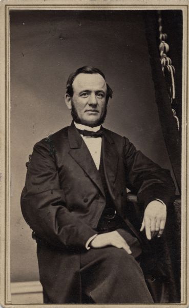 Studio portrait of Elisha Smith sitting. He is wearing a long coat and resting his arm on a table.
