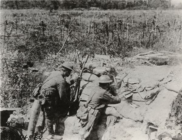 Two soldiers wearing combat uniforms and holding machine guns are in a trench with barbed wire in the background. The men are in a combat zone in France.
