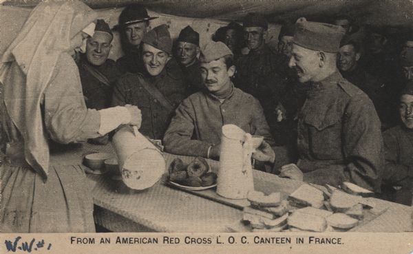 Uniformed soldiers gathered at a counter laden with sandwiches and doughnuts. A woman in a Red Cross uniform at left pours coffee from a carafe.