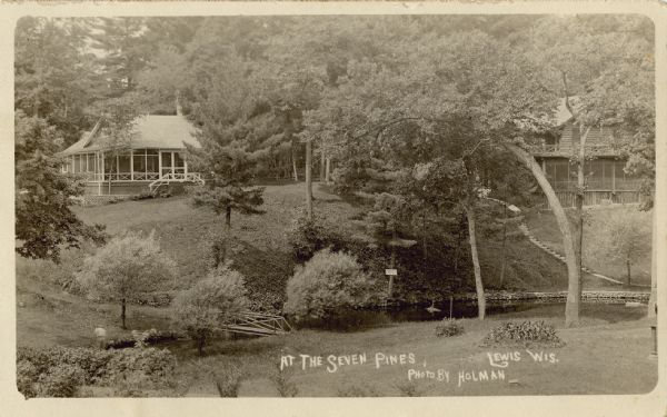 Elevated view across lawn and river toward two buildings on a hill, including the main lodge on the right. A stairway leads down to the river on the right, and a small bridge is crossing it on the left. A man is near the water on the far left. Caption reads: "At the Seven Pines, Lewis, Wis."