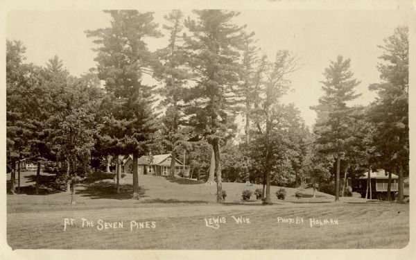 Exterior view of the grounds at the Seven Pines Lodge. Three buildings are in the distance. Caption reads: "At The Seven Pines, Lewis, Wis."