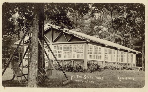 Exterior view of a building with windowed walls. In the foreground is a lawn swing. Caption reads: "At The Seven Pines, Lewis, Wis." Text on back reads: "This is a swimming pool which has a heating plant to warm the water, the interior is all made of tile."