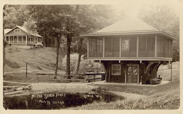 Exterior view of a boathouse, with a canoe on the water nearby. There are two bridges, and abuilding is on hill in the background on the left. Caption reads: "At the Seven Pines, Lewis, Wis."