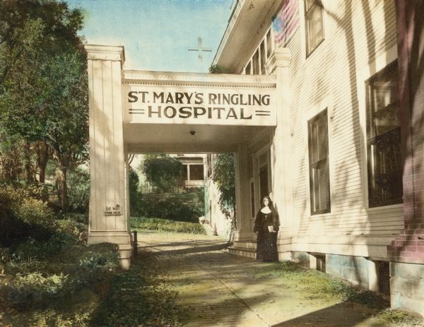 Hand-tinted exterior view of the covered entryway to St. Mary's Ringling Hospital. A sign hanging on the building reads: "Do Not Park Here", and an American flag is on the side of the hospital. A nun is standing near the entryway, and a cross is above the entryway.