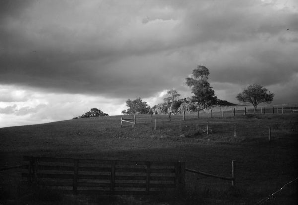 View uphill towards a rocky outcropping on a hilltop near Mount Horeb, with storm clouds gathering in the sky. Fence posts are partway up the hill, and a fence gate is in the foreground.