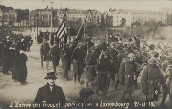 Slightly elevated view of a group of American troops marching into Luxembourg. Several spectators are standing on the street on the left.