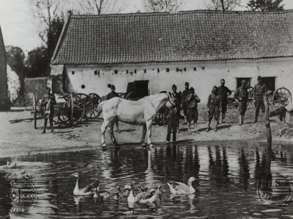 U.S. soldiers are standing in front of a stone farmhouse used for military housing in France. A pond with geese is in the foreground, and a horse is standing at the edge of the pond in the center.