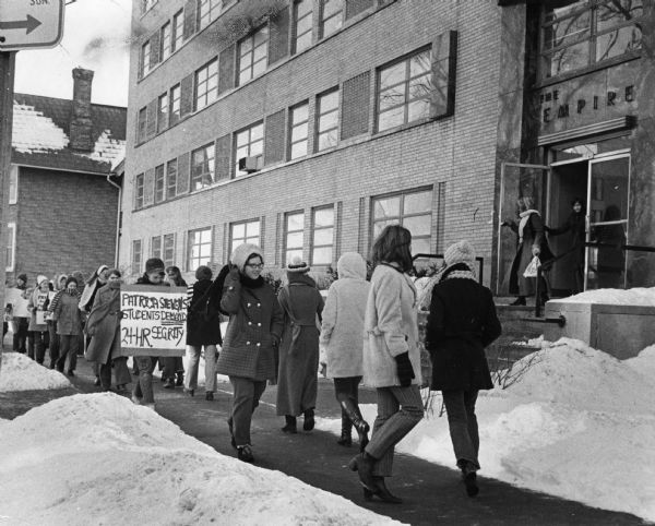 A group of young women are marching on the sidewalk in front of a building with a sign above the entrance that reads: "The Empire." One young woman has a sign that reads "Patricia Stevens Students Demand 24-hr. Security." Caption reads: "Students Seek Security — Sign carrying Patricia Stevens College students demonstrated for 24 hour security protection in front of their dormitory at 1041 E. Knapp St. Monday. The girls organized the protest after a dormitory resident was assaulted inside the building."