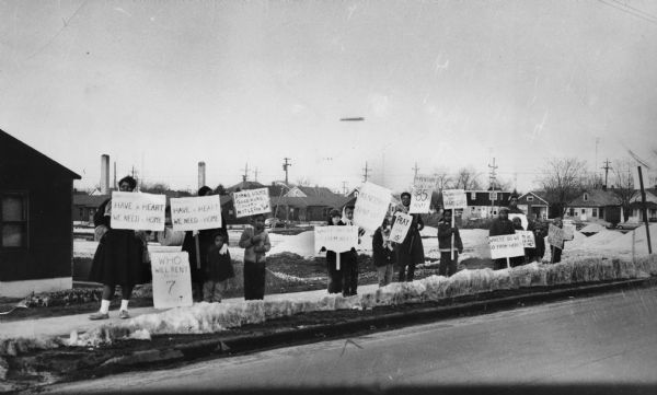 View across street towards several adults and children standing on the sidewalk holding signs, some of which read: "Have a Heart We Need a Home," "Who Will Rent to the 7 of Us?," "Bonnie Hame, Good Home, Colony, Not Leper," "Where Do We Go From Here?," "Fellowships Not Hardships," "Kenosha Tent City," and "Pray With Us." Caption reads: "Members of 26 families at Bonnie Hame protested eviction notices in March, 1961." 
