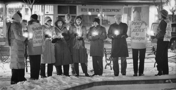 Several people standing together outdoors holding candles. Three people are wearing signs that read: "Waukesha County Peace Council," "PEACE and JUSTICE to Nicaragua," and "PEACE and JUSTICE to El Salvador." Caption reads: "A candlelight vigil, sponsored by the Waukesha County Peace Council, was held at Waukesha's Triangle Park Monday night."