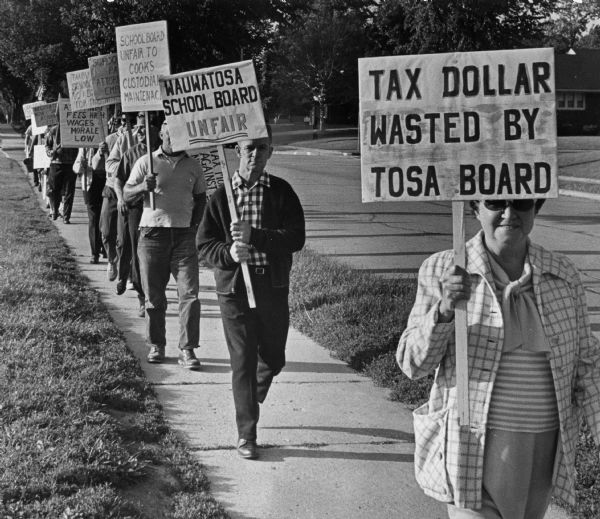 Several people are walking down a sidewalk carrying signs. Some of the signs read: "Tax Dollar Wasted by Tosa Board," "Wauwatosa School Board Unfair," and "School Board Unfair to Cooks, Custodians, Maintenance." Caption reads: "The home of Armand G. Mueller, Wauwatosa school board president, was picketed by public school cafeteria and custodial workers Wednesday. Mueller lives at 7746 Geralayne Circle."