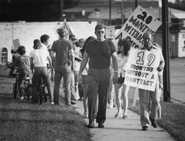 Several people are standing or walking along a sidewalk. Some of the people are holding signs, one of which reads: "19 months without a contract." Caption reads: "Waukesha firemen picketed Tuesday as they protested working without a contract."