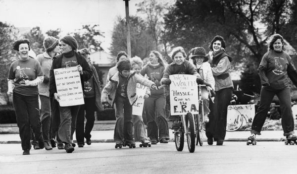 Several women and children are walking, riding bicycles, or on roller skates. Some are carrying signs that read: "Equality of rights under the law shall not be denied or abridged by the United States or any state on account of sex," and "We're worth the 'hassle;' give us the E.R.A." Caption reads: "Skate-A-Way for the ERA — There were skaters, bikers, joggers and walkers who participated in the Skate-A-Way for ERA, sponsored by the Waukesha Chapter of the National Organization for Women. Many carried signs at the start of the fund-raising event as they left the Waukesha County Courthouse." 