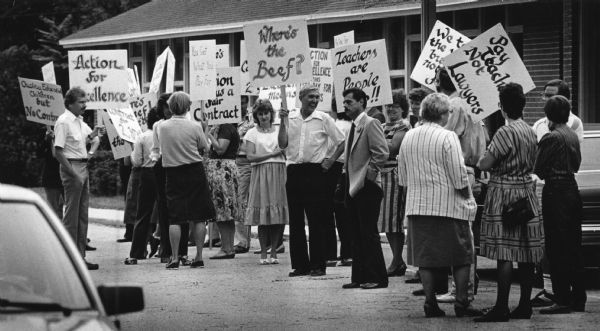 Several men and women are standing outside a building, many holding signs. Among the signs are: "Quality Education for Children but No Control," "Action for Excellence," "You Get What You Pay For," "Where's the Beef?," "Teachers are People [Too]!!," and "Pay Teach[ers] Not Lawyers." Caption reads: "Teachers picketed outside the Franklin School District's offices at 7308 S. North Cape Rd."