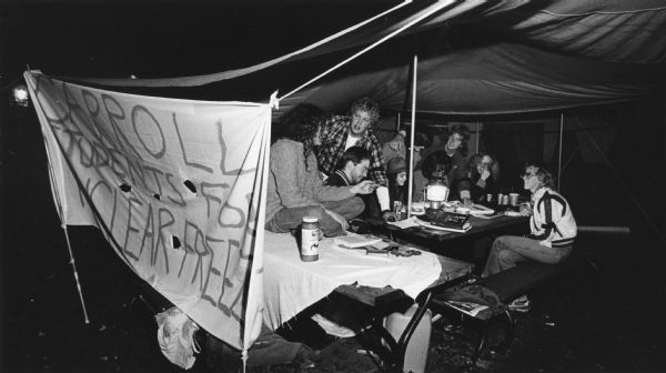 Several college students are sitting on picnic tables under a canopy. They have hung a banner on the side that reads: "Carroll Students for Nuclear Freeze." Caption reads: "Carroll College students holding a vigil for peace and anti-nuke." Carroll College is in front of Old Main Hall northwest of intersection of East and College Avenue.