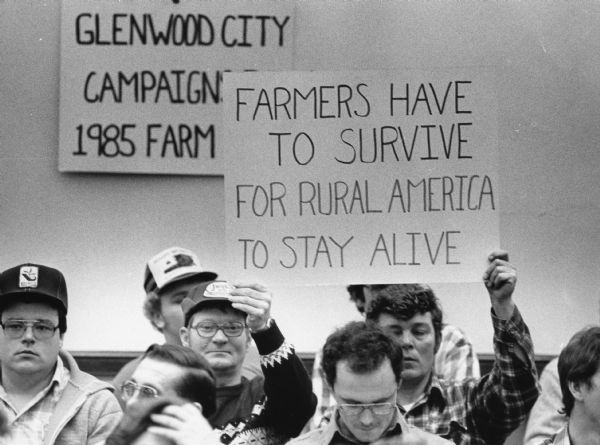 Close-up of two men who are sitting in a group of men and holding a sign which reads: "Farmers Have to Survive for Rural America to Stay Alive." A sign on the wall reads "Glenwood City Campaigns [?] 1985 Farm [?]." Caption reads: "Hundreds of farmers and supporters demonstrate in an effort to call attention to what some consider the worst farm conditions since the Great Depression."