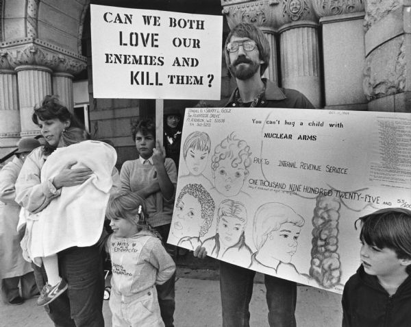 A man, a woman, and four children are standing in front of building with an arch and columns. They are holding signs that read: "Can We Both LOVE Our Enemies and KILL Them?" and a sign made somewhat like a check with the title: "You can't hug a child with NUCLEAR ARMS." Caption reads: Checking In — Sherry and Randy Golz had a check for the Internal Revenue Service as their family demonstrated Wednesday outside the IRS offices at 517 E. Wisconsin Ave. From left were Sherry (holding Rebecca); Dawn (with sign) Stephanie, Randy, and David."