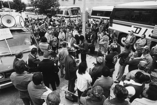 A crowd of people are gathered around a man standing center left and speaking into a microphone attached to a vehicle which has megaphones attached to its roof. In the center is a man holding a sign, the words of which are not completely visible but which probably read "On Strike." Caption reads: "More than 800 people listened to speeches Friday in the 600 block of N. 24th St. before boarding buses for a ride to Washington, D.C., for a 'Solidarity Day' rally."